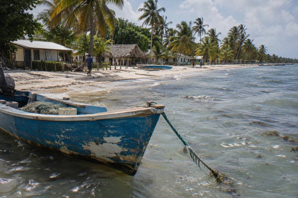 A wooden fishing boat moored in shallow clear water beside a sandy shoreline lined with palms