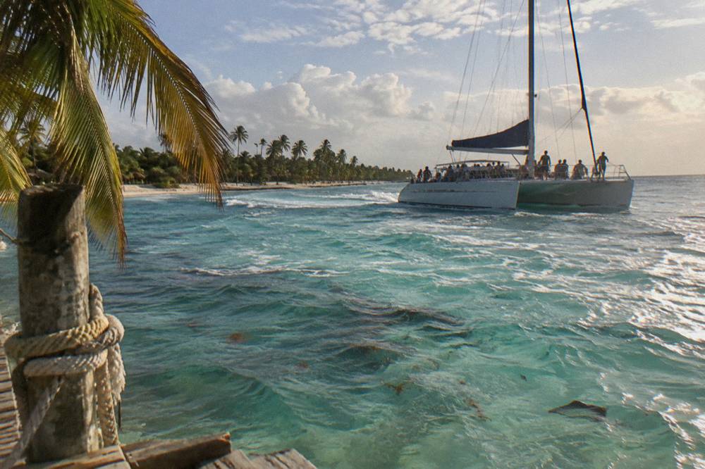 A large catamaran with passengers aboard sailing across shallow turquoise water near an island