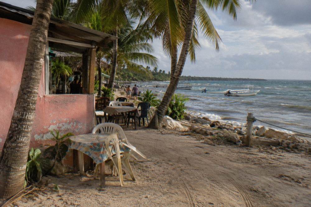Outdoor restaurant tables under palm trees beside a village street in late afternoon