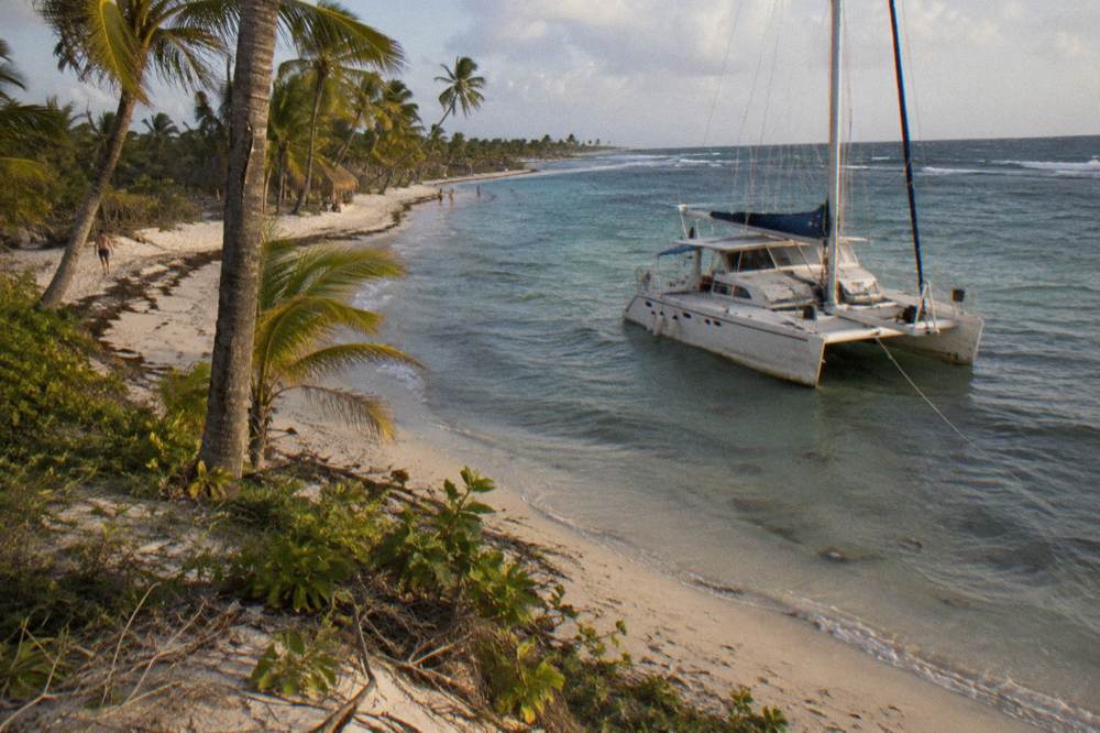 Catamaran moored in clear shallow water beside a sandy spit with scattered palms
