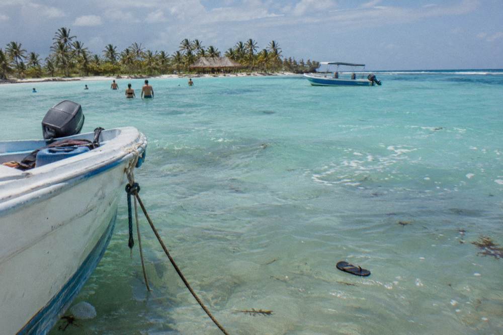 Boats anchored on a shallow sandbar where people stand in clear waist-deep turquoise water far from any shore