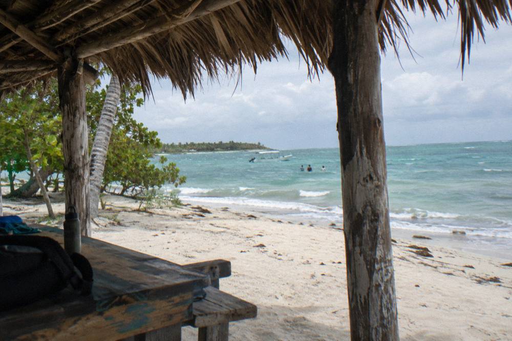 A thatched palm roof shading white sand with calm green blue water beyond
