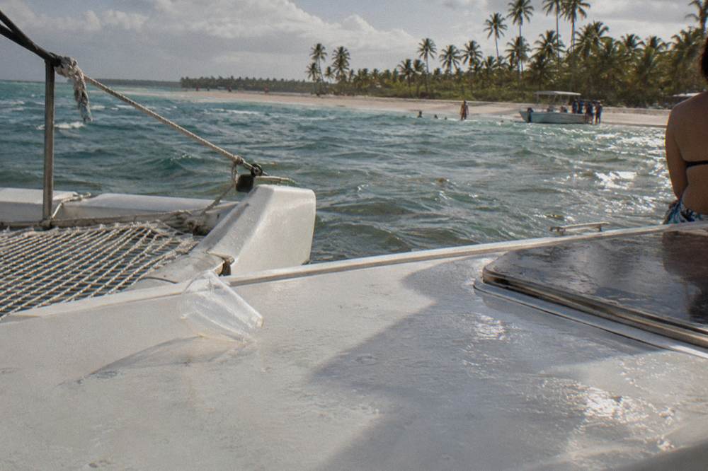 A catamaran moored in shallow clear water near a sandbar with distant palm trees