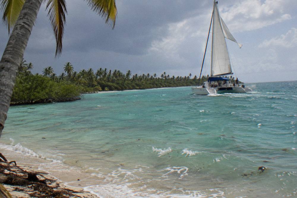 Catamaran sailing on calm turquoise water toward a green island shoreline