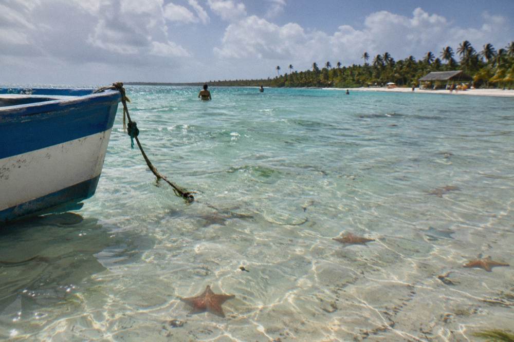 Shallow turquoise water over a sandbar with distant palm-lined shoreline