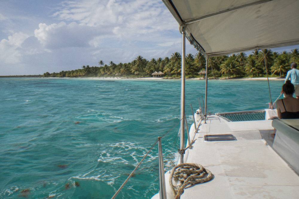 A covered catamaran cruising across shallow turquoise water toward a tree-lined island