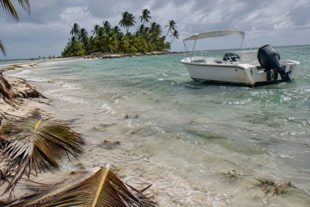 A white speedboat anchored in shallow clear water beside a sandbar dotted with palm fronds