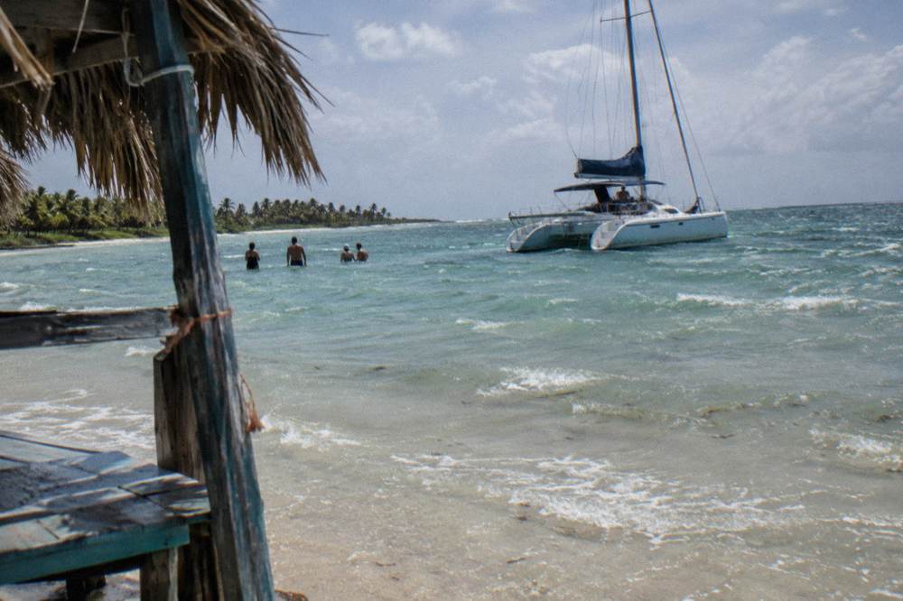 A shallow sandbar with bathers standing in clear water beside a moored catamaran