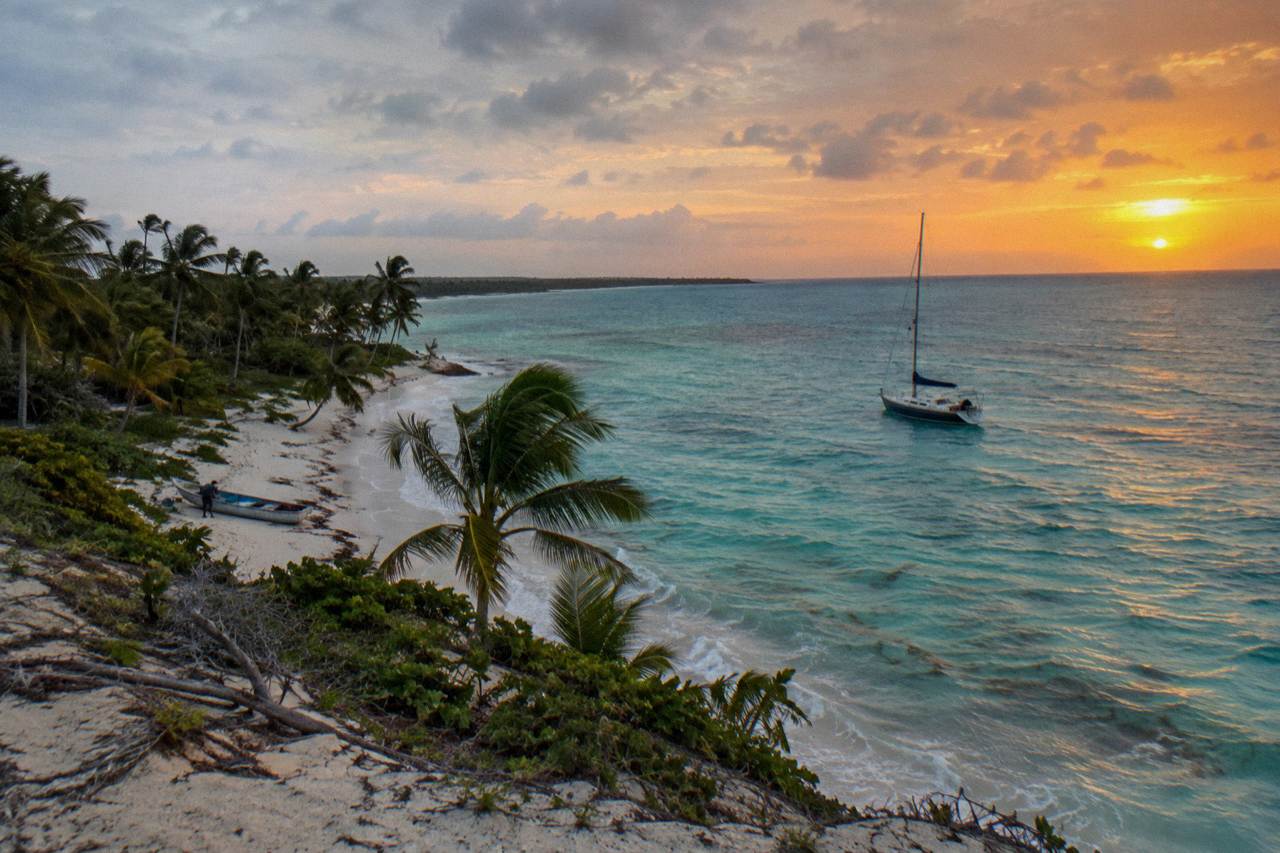 Anchored sailboats resting on calm turquoise shallows beside a palm-lined tropical island shoreline at late afternoon