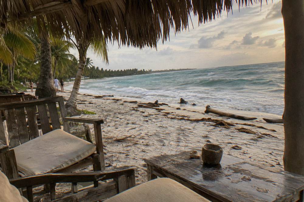 Shaded beach lounge area with wooden furniture under palm trees on white sand