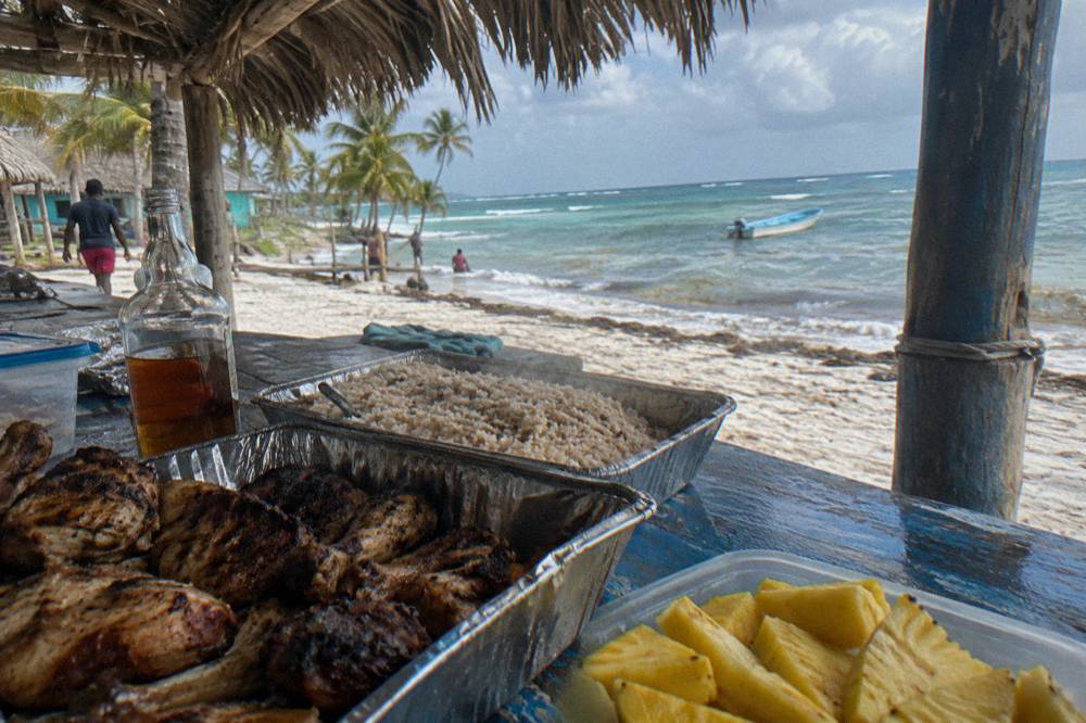 Buffet lunch table with grilled food and tropical fruit under a palm-thatch shelter