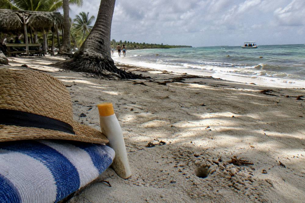 Beach towel, hat and sunscreen arranged on white sand under palm shade