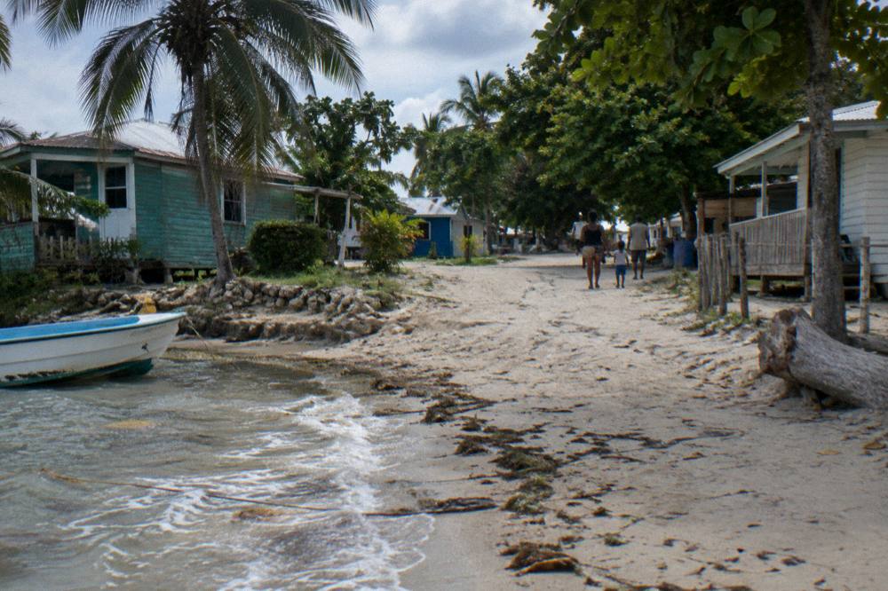 Families walking a sandy village path between wooden houses near a beach landing