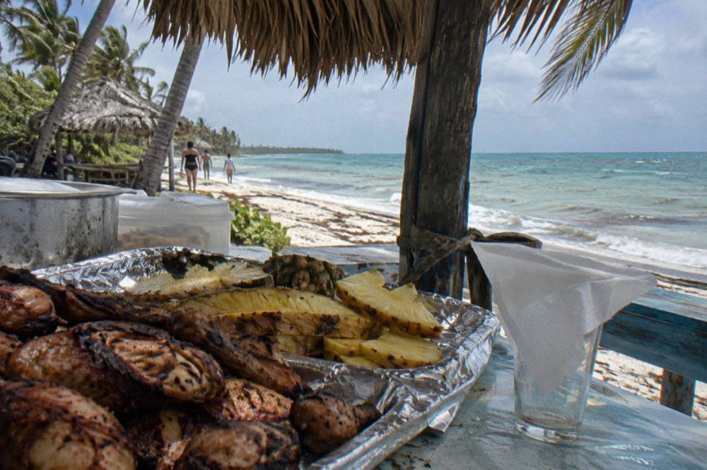 A shaded beach buffet table with grilled food and tropical fruit under palm fronds