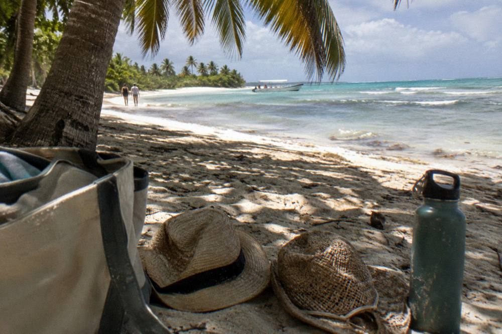 A beach bag, sun hats and a reusable water bottle on sand under palm shade