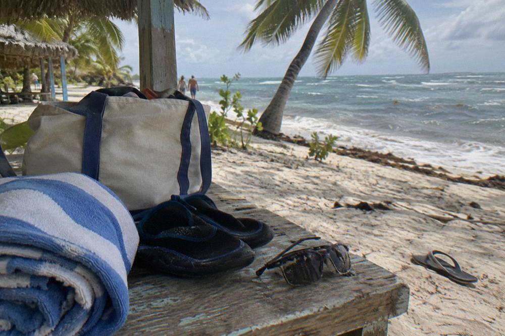 A canvas tote bag with a hat, sunglasses and sunscreen resting on a wooden bench beside a beach towel